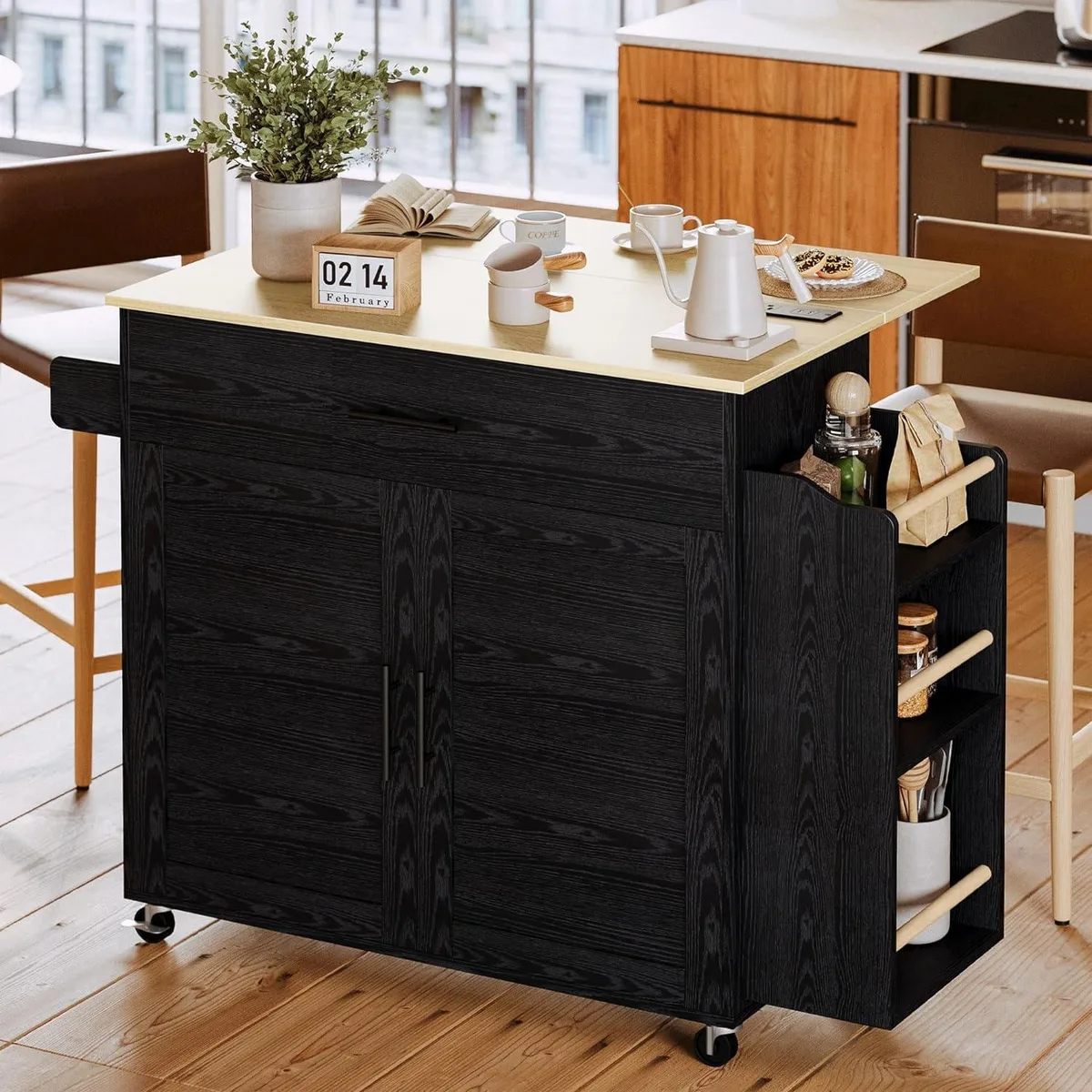 Black rectangular kitchen island on four wheels with drop leaf extended, open spice rack, cabinet door, drawer and charging station