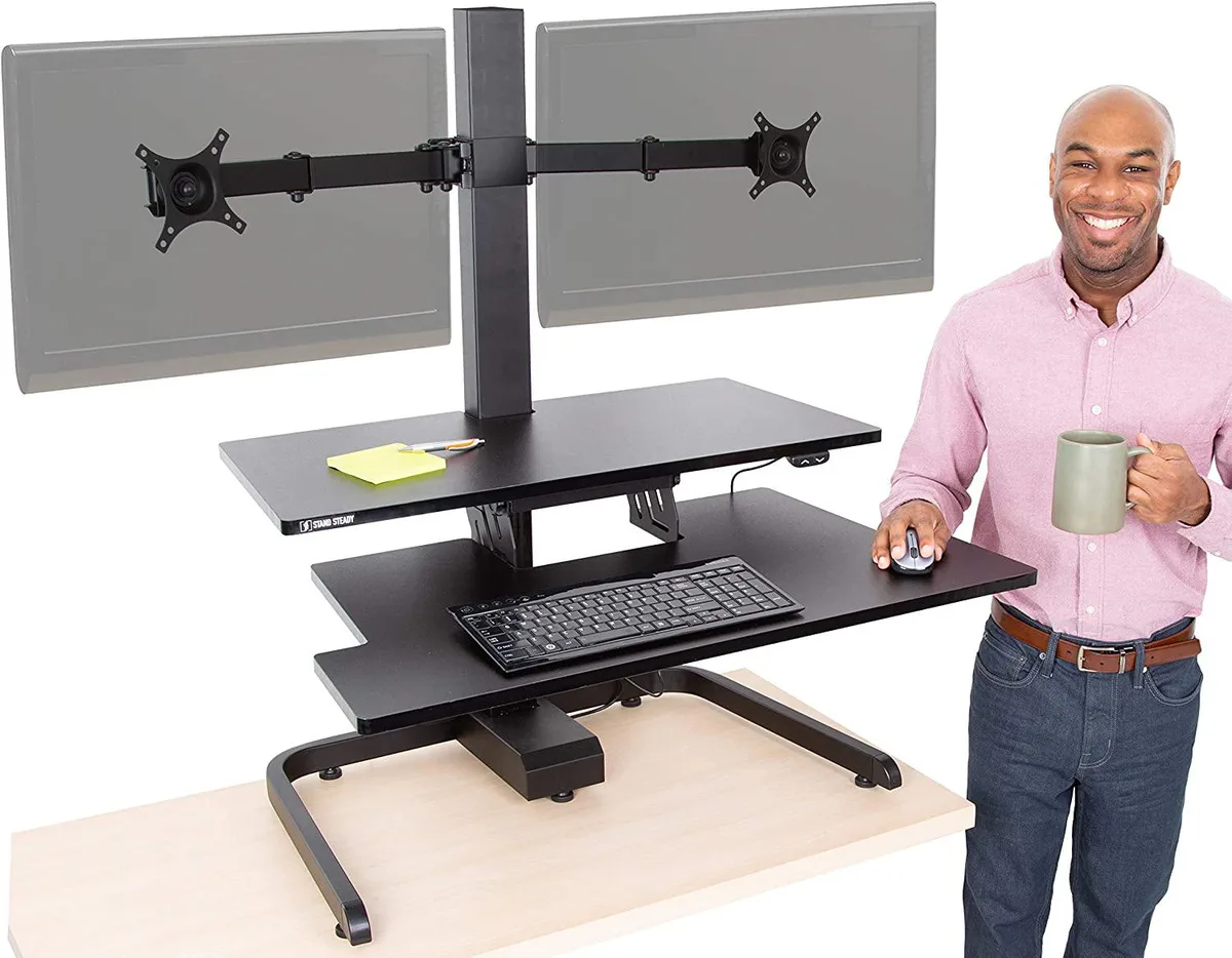Black electric standing desk converter with dual monitor arms and keyboard tray on desktop
