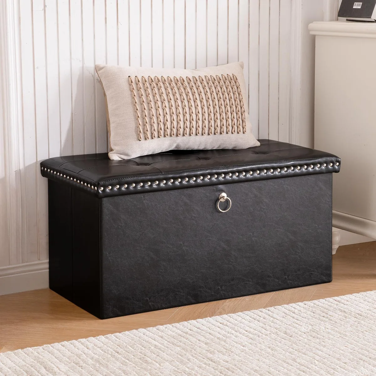 Rectangular black faux-leather storage ottoman bench, button-tufted lid with rivet trim, folded flat showing MDF base