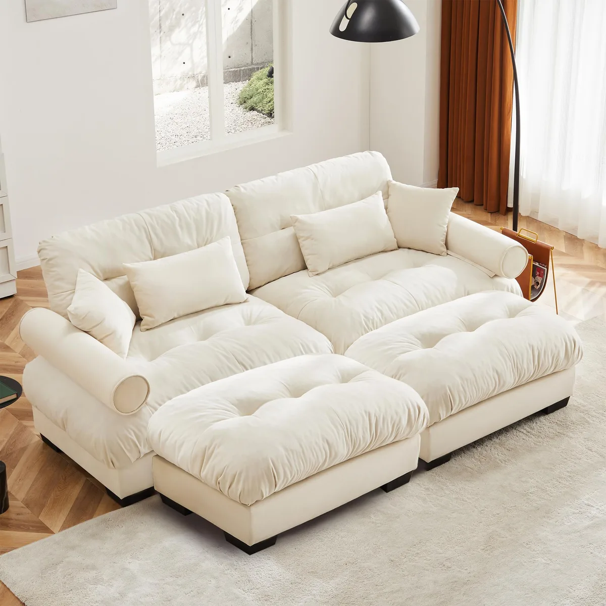 Cream velvet two-seater sofa with matching ottoman, round bolster armrests, and decorative pillows