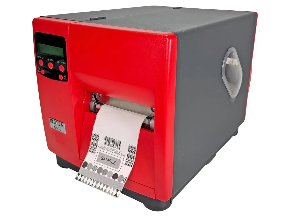 Rectangular industrial barcode printer in light grey, front panel with display and controls, label roll compartment visible